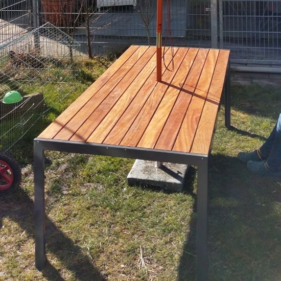 Gartentisch mit hellbraunen Holzlatten auf schwarzem Metallgestell auf Rasen vor Drahtzaun, Sonnenlicht wirft Schatten, rechts teilweise eine Person mit Jeans und Schuhen sichtbar