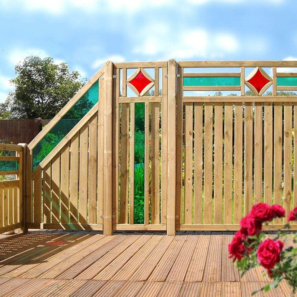 Holzzaun Monticiano mit senkrechten Lamellen, farbigen Glaseinsätzen in Rot und Grün im oberen Bereich, auf Holzterrasse, im Vordergrund rote Rosen, im Hintergrund Garten und blauer Himmel