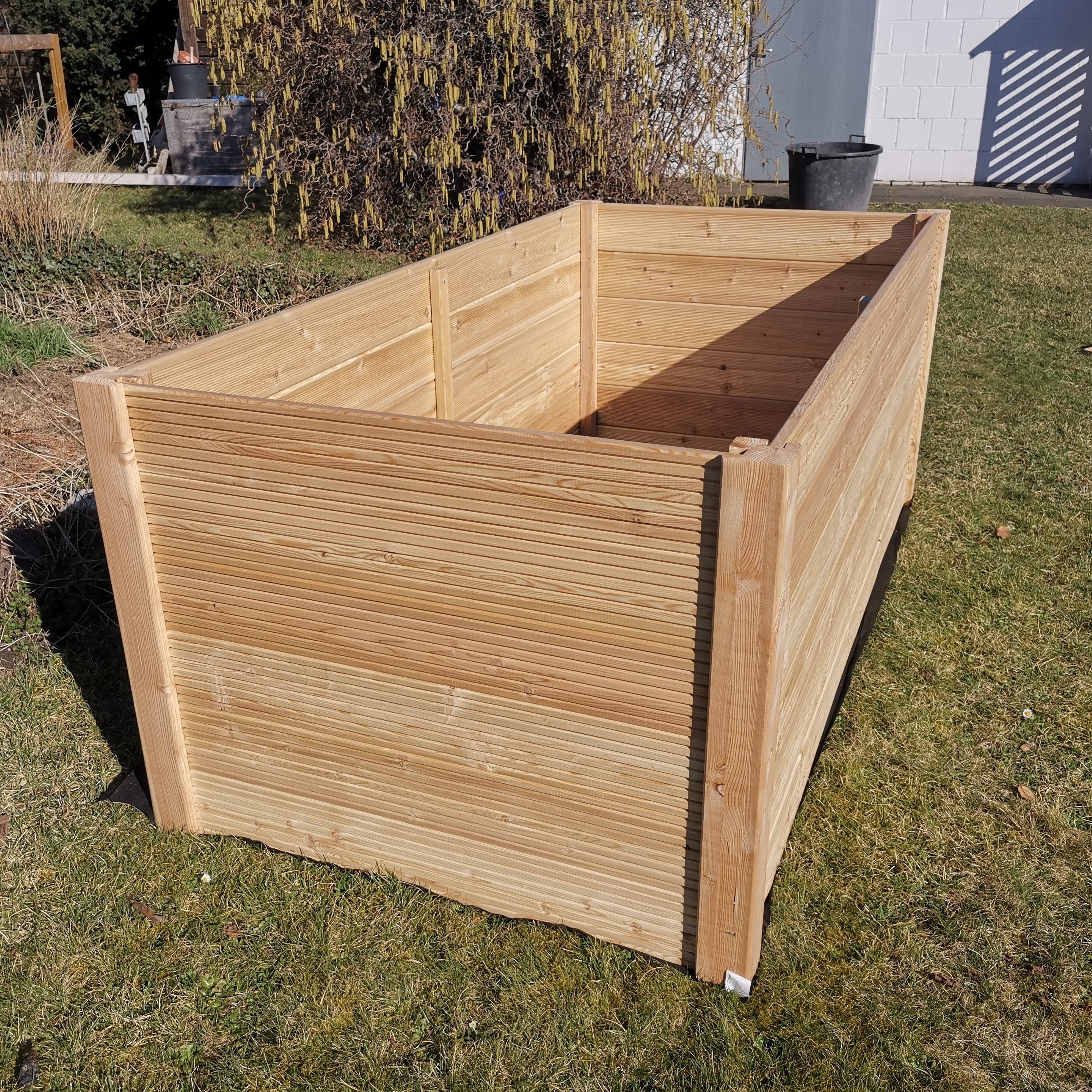 Rechteckiges Hochbeet aus 24 mm starkem Lärchenholz, außen grob geriffelt, innen glatt, auf grünem Rasen im Garten aufgestellt, Vorder- und Seitenansicht bei Sonnenschein