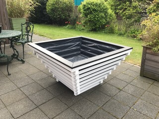 Quadratischer, nach unten verjüngter weiß gestrichener Holzkübel mit schwarzer Innenfolie auf gepflasterter Terrasse; links grüner Metall-Gartentisch und -stuhl, im Hintergrund Rasen und Büsche