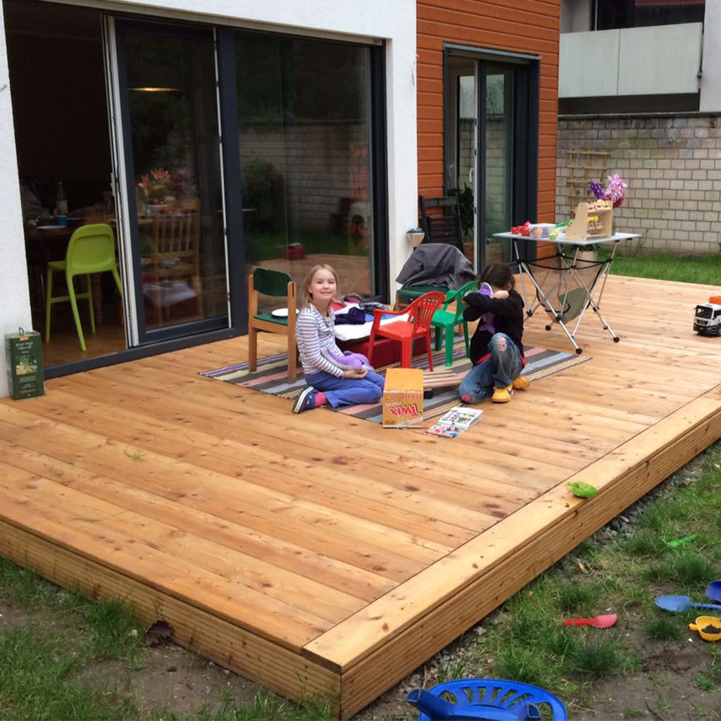 Zwei Kinder sitzen auf einer Holzterrasse vor einem modernen Haus mit großen Schiebetüren; bunte Kindermöbel, Spielzeug auf einem Teppich, ein Klapp­tisch mit Bastelmaterialien und verstreute Spielsachen im angrenzenden Gras