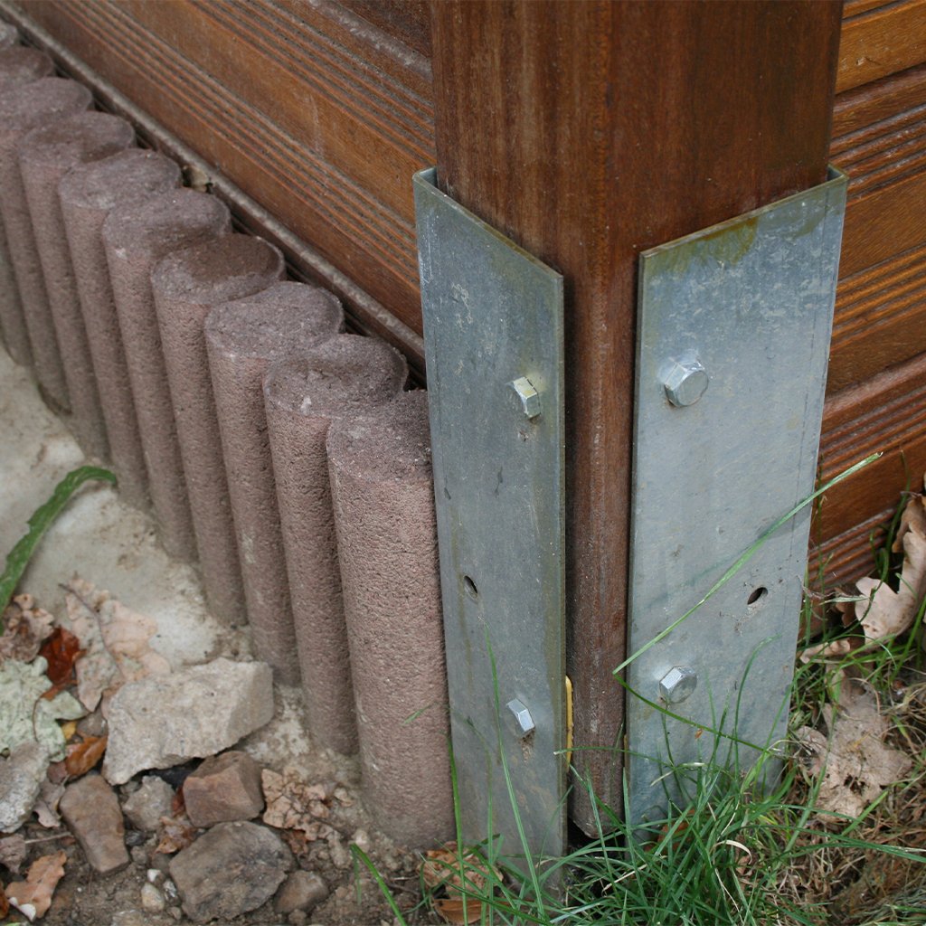 Winkelanker aus Metall zum Einbetonieren, befestigt an einem Holzpfosten, teils umgeben von Erde, Steinen, Gras und Betoneinfassung; Schrauben sichern den Anker am Holzpfosten