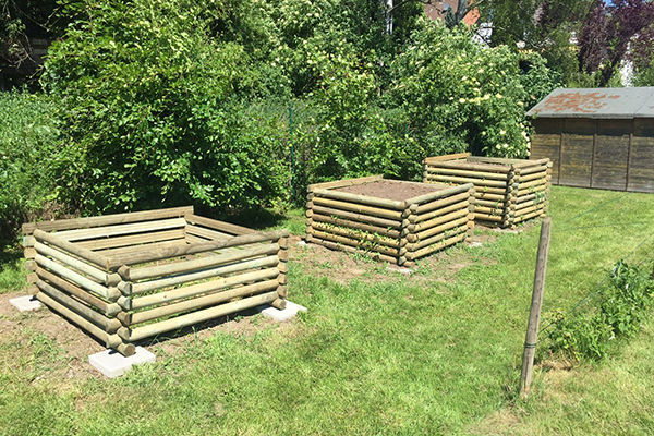 Drei quadratische Hochbeete aus übereinander geschichteten Rundhölzern auf Rasen, im Hintergrund Büsche und ein kleiner Holzschuppen bei sonnigem Wetter