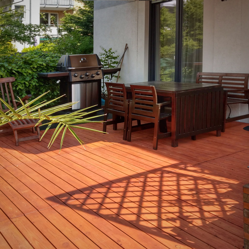 Terrassenbereich mit rotem Holzdeck, dunklem Holztisch und Stühlen, Edelstahl-Gasgrill, Palmenblatt im Vordergrund, Glasschiebetür und grüner Hecke im Hintergrund, dekorativer Schatten auf dem Boden