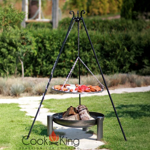 Dreibeiniger Kochständer mit hängendem Grillrost, auf dem Würstchen und geschnittenes Gemüse liegen; darunter eine Feuerschale mit Holzscheiten, auf Rasen im Garten mit Hecke und Pflanzen im Hintergrund