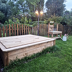 Holz-Sandkasten mit geschlossenem Deckel auf grünem Rasen vor einem Lattenzaun, daneben eine kleine hölzerne Spielküche/Werkbank mit Spielzeug und Gießkanne, Laternenbeleuchtung und Bäume im Hintergrund
