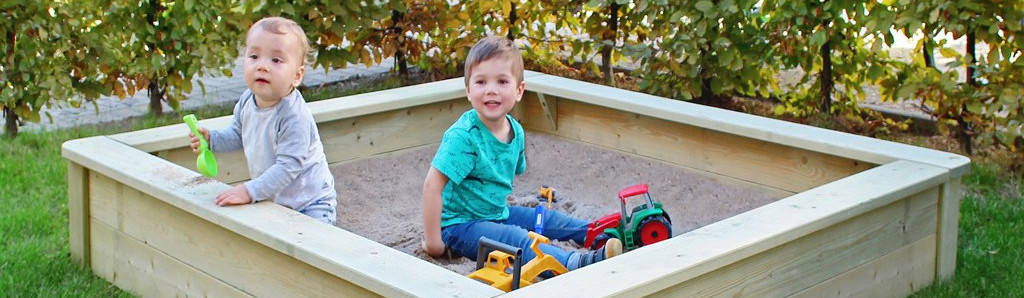 Zwei Kleinkinder sitzen in einem rechteckigen Holzsandkasten im Garten, umgeben von Rasen und Hecke; eines mit grauem Shirt hält eine grüne Sandschaufel, das andere in türkisfarbenem Shirt lächelt und spielt mit gelbem Bagger- und rotem Traktor-Spielzeug