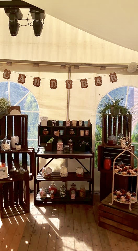 Zelt-Candybar mit Girlande CANDYBAR, hölzernen Kisten und Regalen mit Bonbongläsern, Schalen und süßen Leckereien, zwei Bogenfenstern im Hintergrund, Holzboden und Bühnenbeleuchtung an der Decke