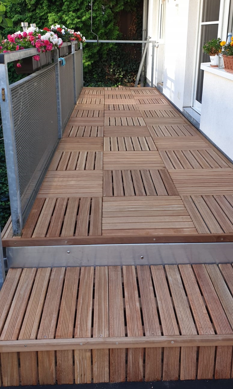 Balkon mit neu verlegten Holzfliesen im Schachbrettmuster, Metallgeländer links mit Blumenkästen (rosa und weiße Blumen), weiße Hauswand und Tür rechts, Stufe im Vordergrund und dichte grüne Hecke am Ende