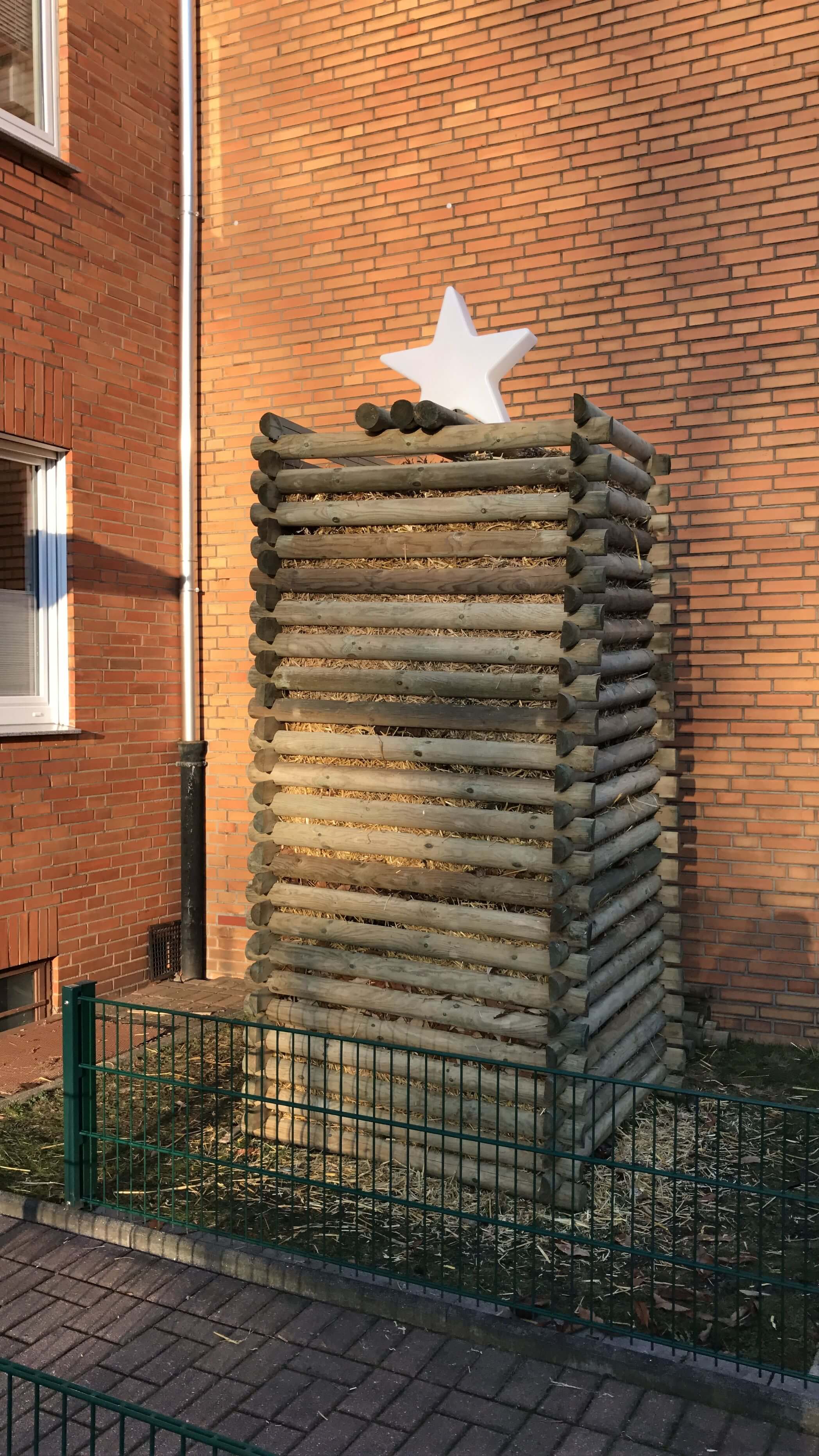 Hoher quadratischer Turm aus gestapelten Rundhölzern gefüllt mit Stroh, obenauf eine weiße Stern-Dekoration vor roter Backsteinfassade, grüner Metallzaun im Vordergrund