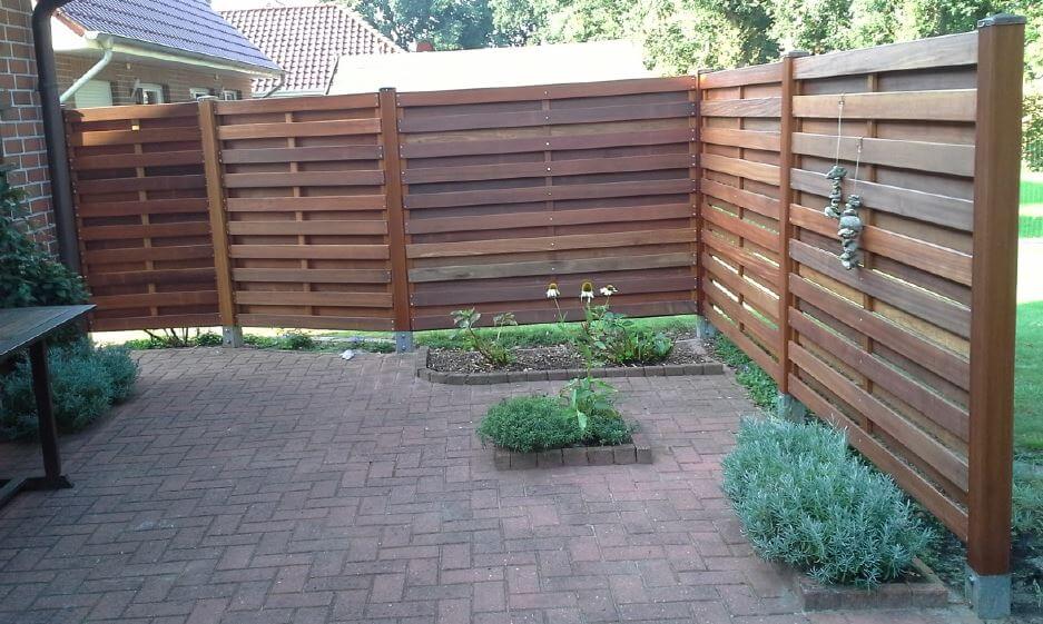 Terrasse mit rotbraunem, horizontalem Holzsichtschutz in Eckform, Backsteinpflaster, kleine Beete mit Blumen und Kräutern in der Mitte und rechts, hängende Stein-Dekoration am rechten Pfosten, Hauswand links und Rasen im Hintergrund