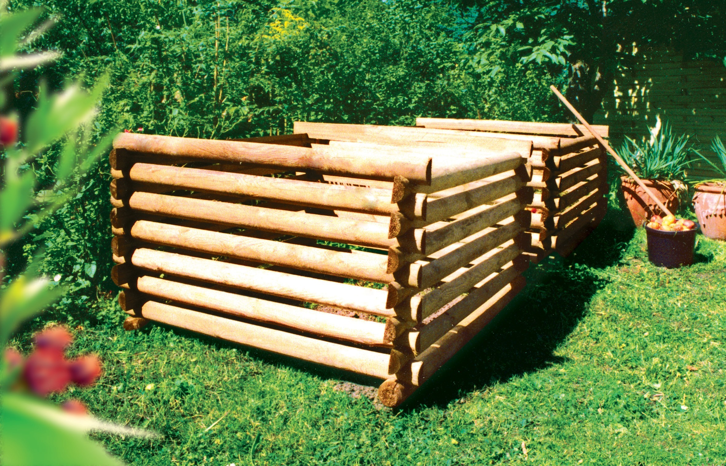 Gestapelte, ineinandergreifende runde Holzbalken auf einer Wiese, rechts eine Stange, Pflanzkübel und ein Eimer mit Äpfeln, im Hintergrund dichtes grünes Gebüsch und links unscharfe Blätter im Vordergrund