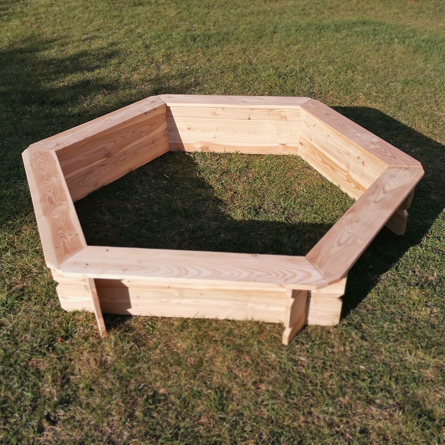 Sechseckiger Sandkasten aus Lärchenholz mit einem Durchmesser von 174 cm, aufgebaut auf einer Wiese, Sicht von schräg oben, unbehandelte Holzstruktur deutlich erkennbar