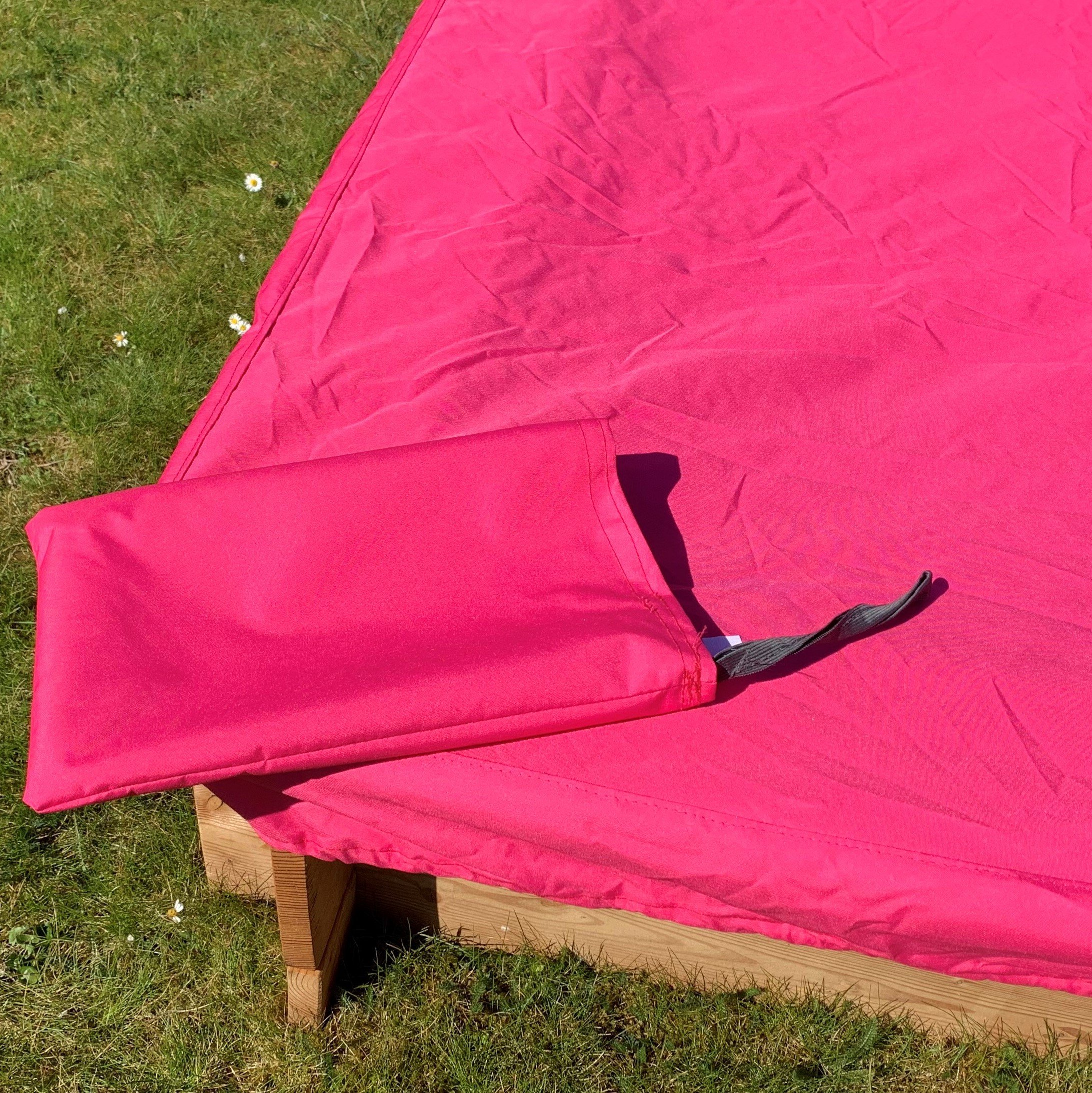 Rote Abdeckplane auf einem Holzsandkasten im Garten, eine Ecke der Plane ist umgeschlagen und zeigt einen angenähten Befestigungsriemen, grüner Rasen mit Gänseblümchen im Hintergrund