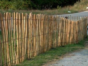 Haselnuss Staketenzaun, naturbelassen, 120 cm hoch, aus senkrechten, unregelmäßig geformten Holzlatten und waagerechten Drähten, steht auf einer Wiese am Straßenrand