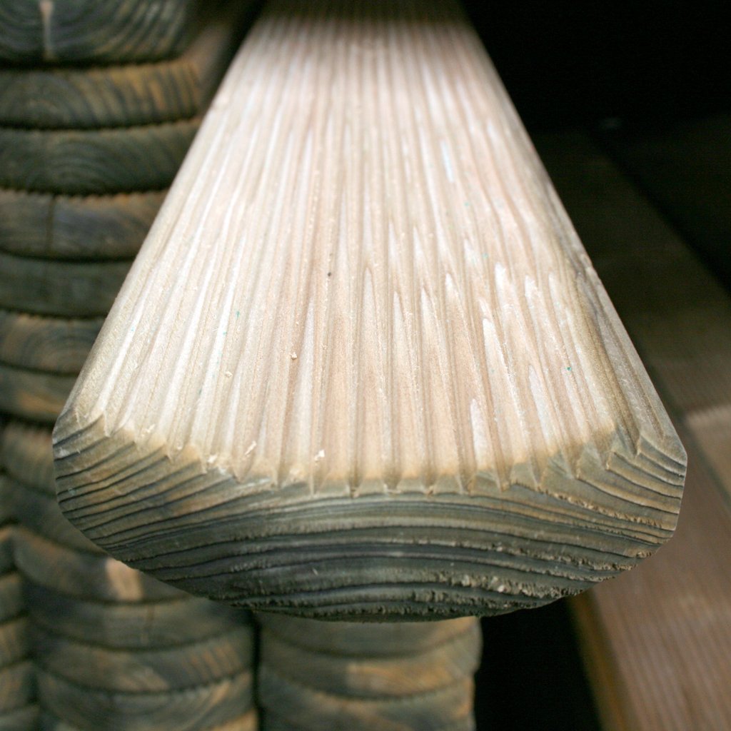 Nahaufnahme eines kesseldruckimprägnierten Zaunbretts Mosel mit Rundkopf und geriffelter Oberfläche, Maße 22 x 95 mm, Stapel weiterer gleichartiger Bretter im Hintergrund