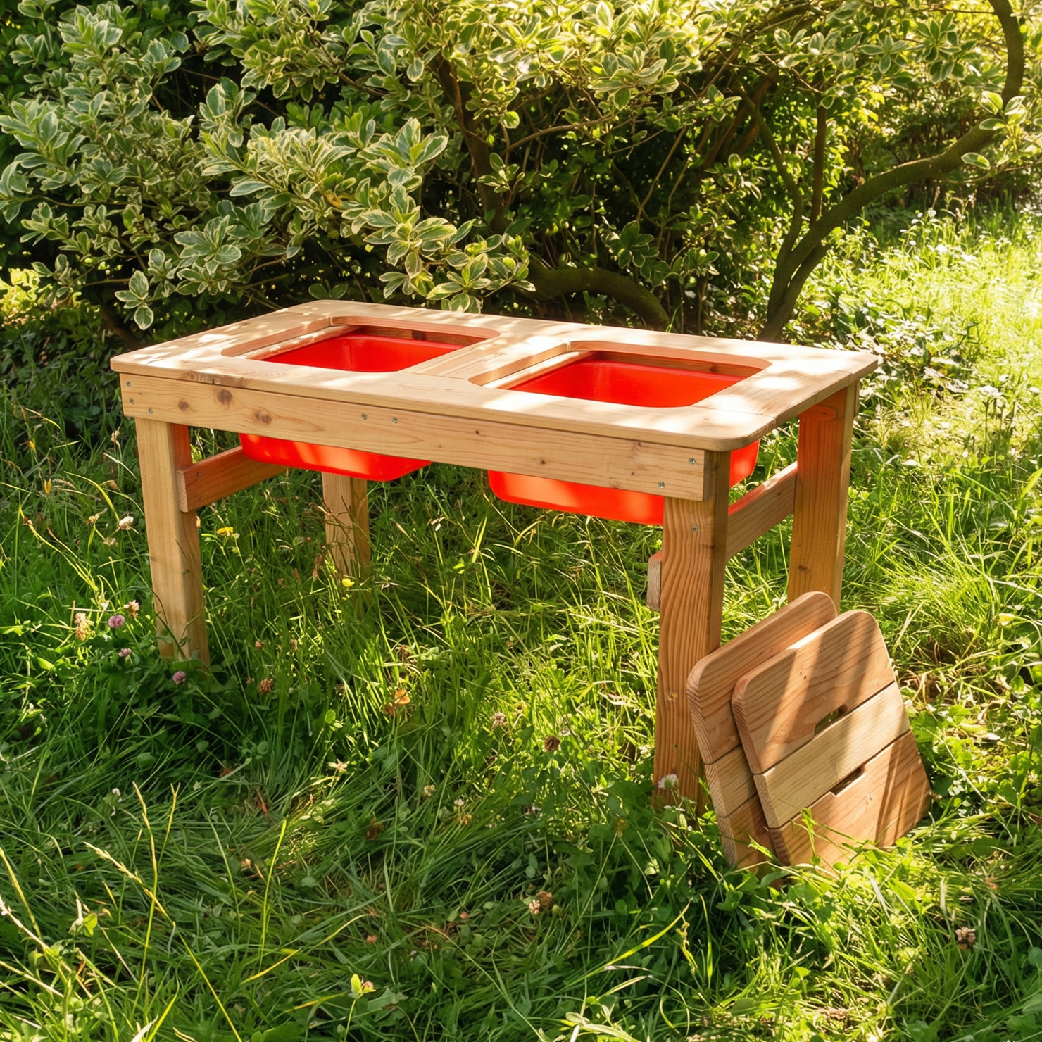 Spieltisch Wassermax aus Lärche (100×50×60 cm) mit zwei roten Kunststoff-Einsätzen, stabiles Holzgestell und aufgestellte Holzbank, steht im Gras vor einem dichten Strauch