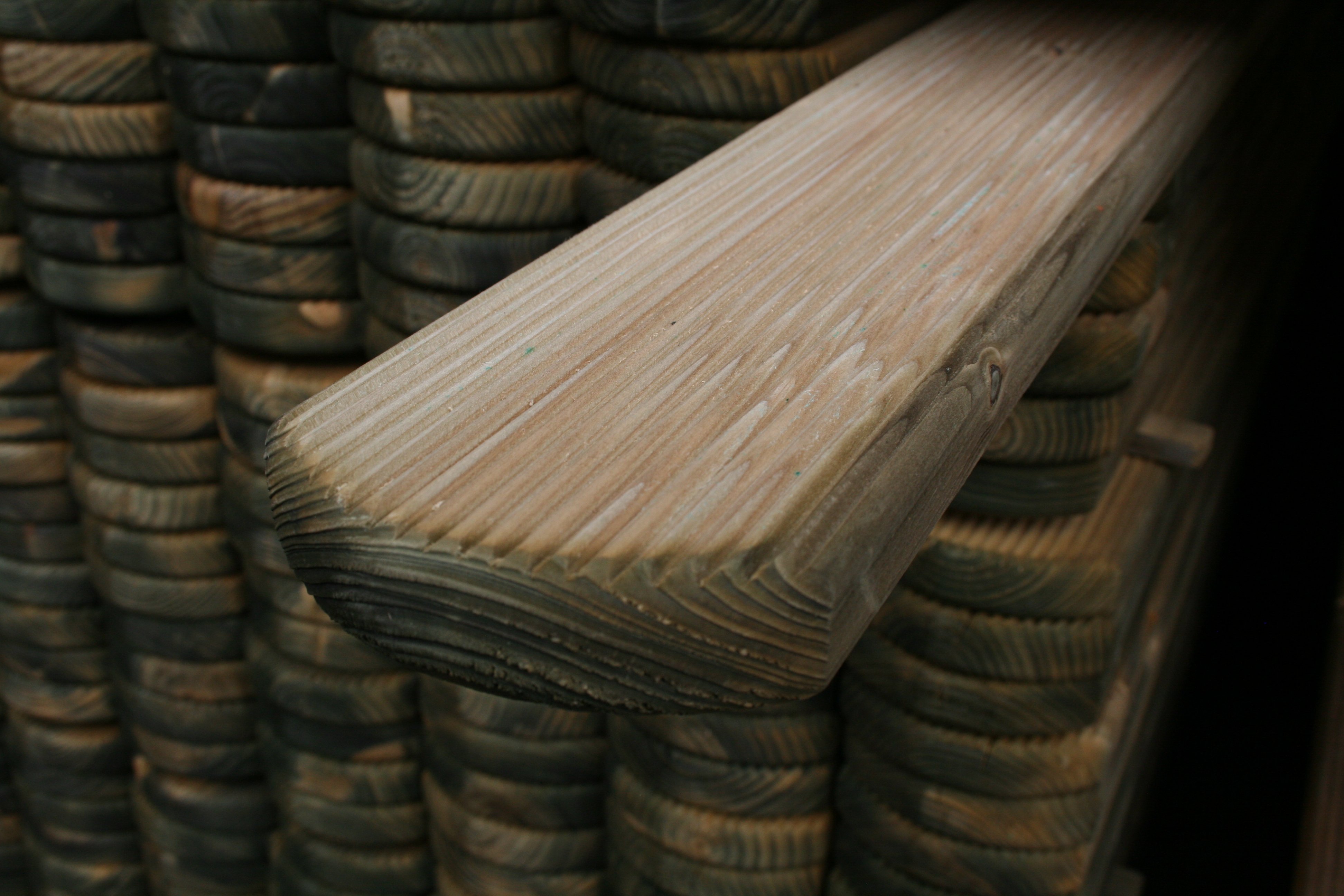 Detailansicht eines geriffelten, kesseldruckimprägnierten Zaunbretts Mosel mit abgerundetem Kopf, Stapel von ähnlichen Holzbrettern im Hintergrund, Maße 22 x 95 mm