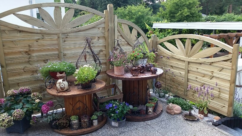 Zaun Verona Ornamentzaun aus imprägnierter Kiefer mit dekorativem Sonnenmuster, als Sichtschutz auf Terrasse, davor bepflanzte Kabeltrommeln, verschiedene Blumentöpfe und Gartenaccessoires