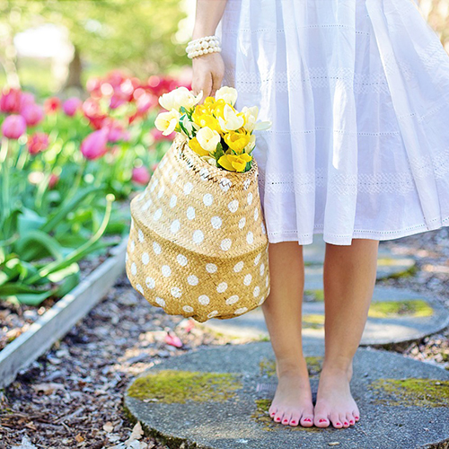 Barfüßige Frau im weißen Sommerkleid steht auf moosigem Steinweg, trägt Perlenarmband und hält gepunkteten Strohkorb voller gelber und weißer Tulpen, im Hintergrund blühende pinke Tulpen entlang eines Gartenwegs