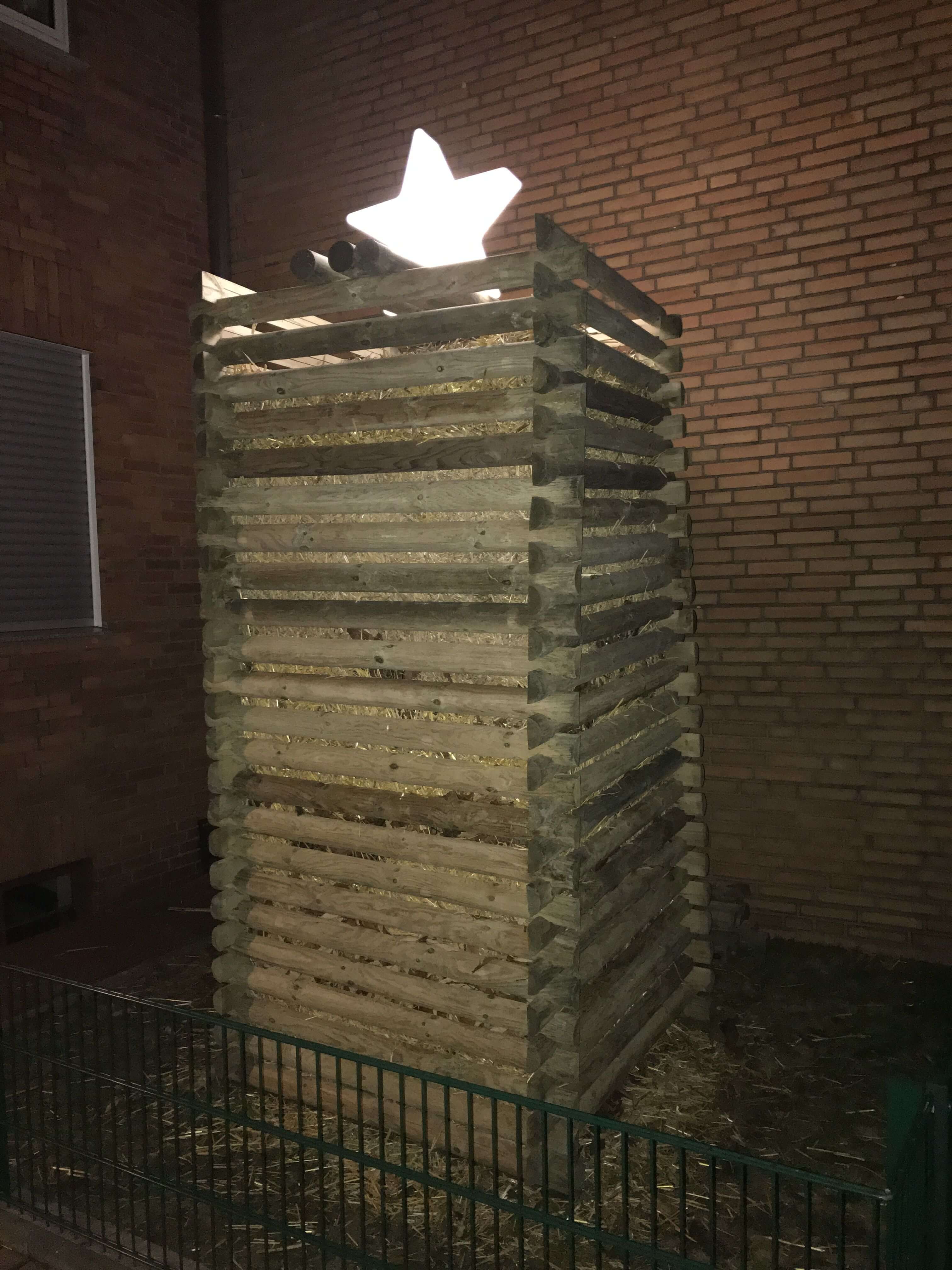 Gestapelte Holzkonstruktion mit Strohfüllung und leuchtendem Stern oben, grüner Metallzaun im Vordergrund, rote Backsteinwand im Hintergrund bei Nacht