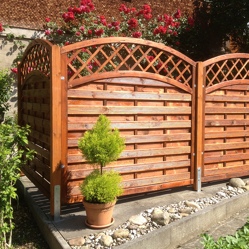 Sonniger Garten mit geschwungenem Holzsichtschutz mit Rautenmuster, davor Terrakotta-Topf mit zweistufiger Konifere, Kies- und Steinrand und im Hintergrund blühende rote Rosen