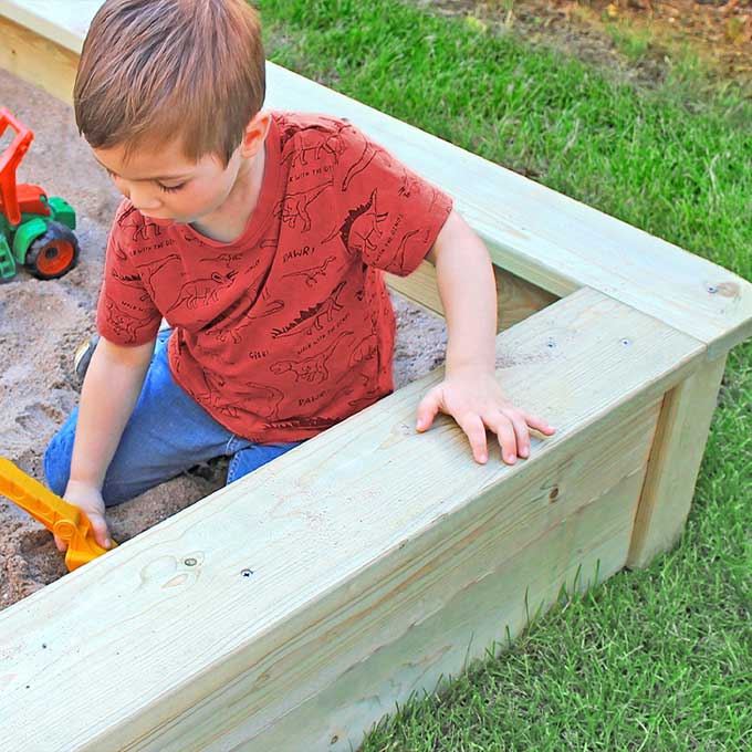 Kleiner Junge sitzt im Holzsandkasten auf dem Rasen, trägt rotes Dinosaurier-T-Shirt und gräbt mit gelber Schaufel; grüner Spielzeugtraktor liegt daneben.