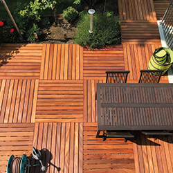 Terrassenholz in quadratischem Verlegemuster mit warmen Holzfarben; dunkelbrauner Esstisch mit Stühlen, grüner Pflanzkübel, Gartenschlauchrolle und bepflanztes Beet am Rand; Sonnenlicht erzeugt Schattenmuster