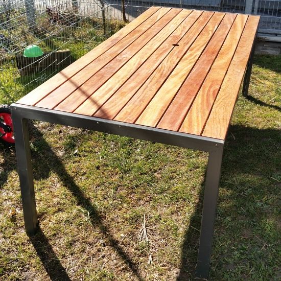 Rechteckiger Gartentisch mit hellen Holzlatten und grauem Metallgestell auf Gras, Sonnenlicht wirft Schatten, im Hintergrund ein Maschendrahtzaun