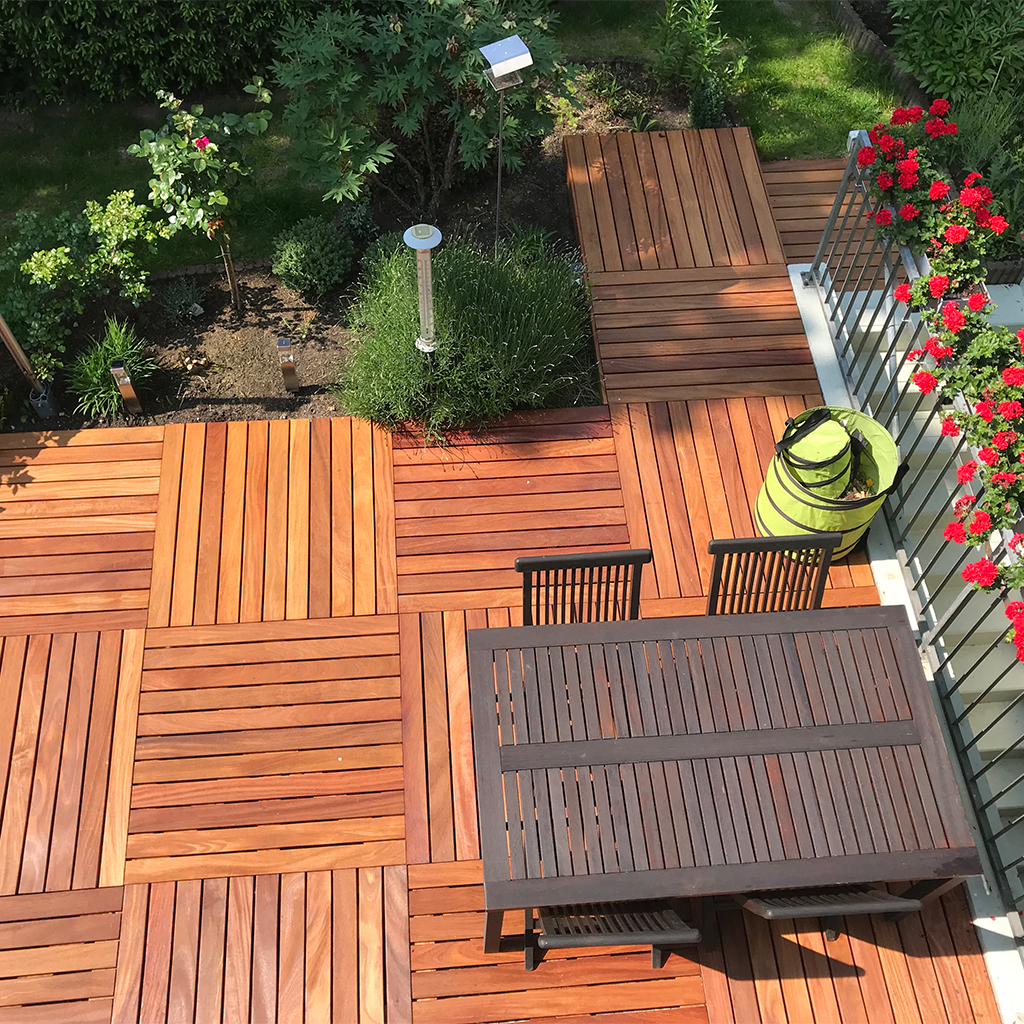 Vogelperspektive auf sonnige Holzterrasse mit quadratischen Holzfliesen, dunklem Gartentisch und Stühlen, rechte Balkonbrüstung mit roten Pelargonien, grüner Falt-Gartensack, oben angrenzendes Beet mit Sträuchern und Gartenlampen
