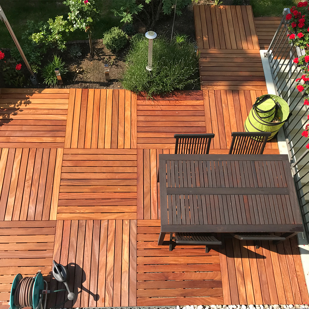 Draufsicht auf Terrasse mit quadratischen Holzfliesen in wechselnder Maserung, dunkler Holztisch und zwei Stühlen, rechts Balkon mit roten Blumen, oben Beet mit Sträuchern und Laterne, unten Gartenschlauch auf Rolle