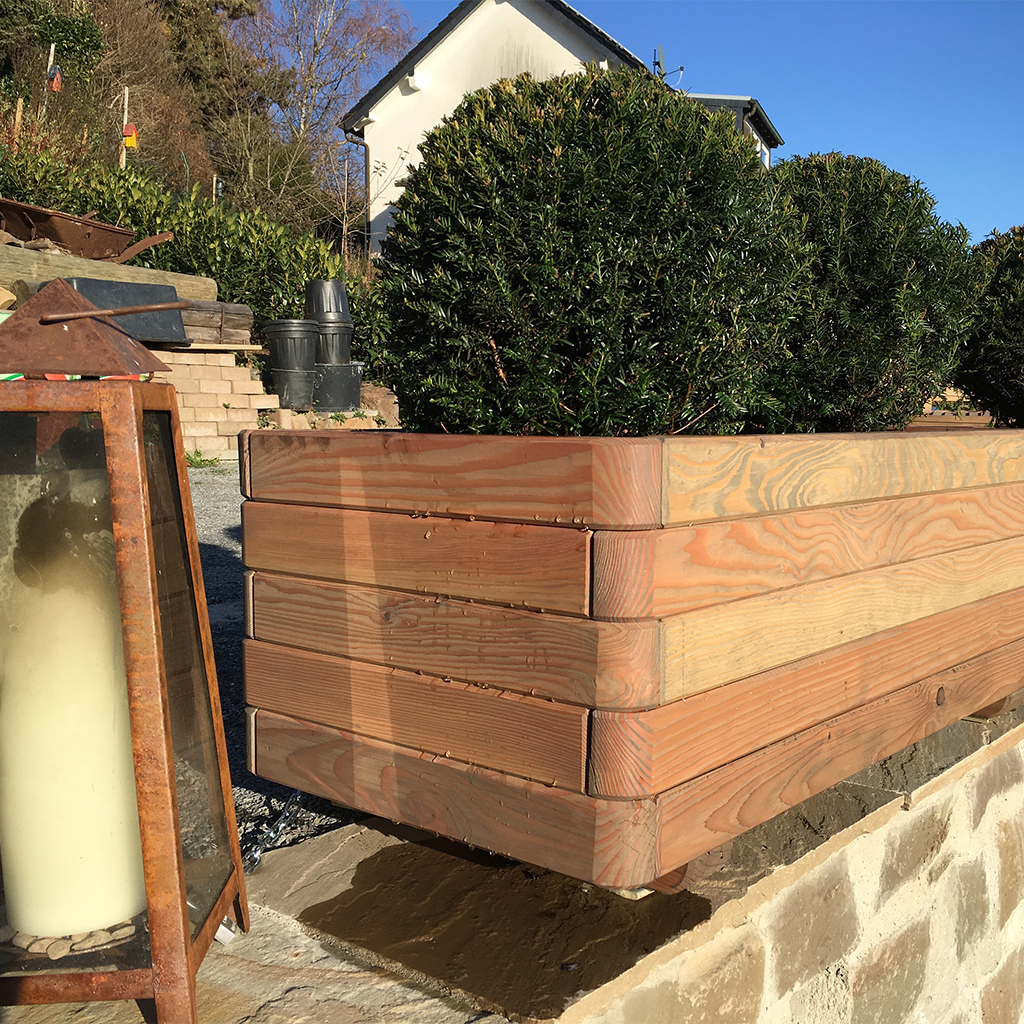 Holz-Pflanzkasten mit kugelförmigen Buchsbaumsträuchern auf Steinmauer, geschichtete Nadelholzlatten, links rostige Laterne mit großer weißer Kerze, im Hintergrund weißes Haus und blauer Himmel