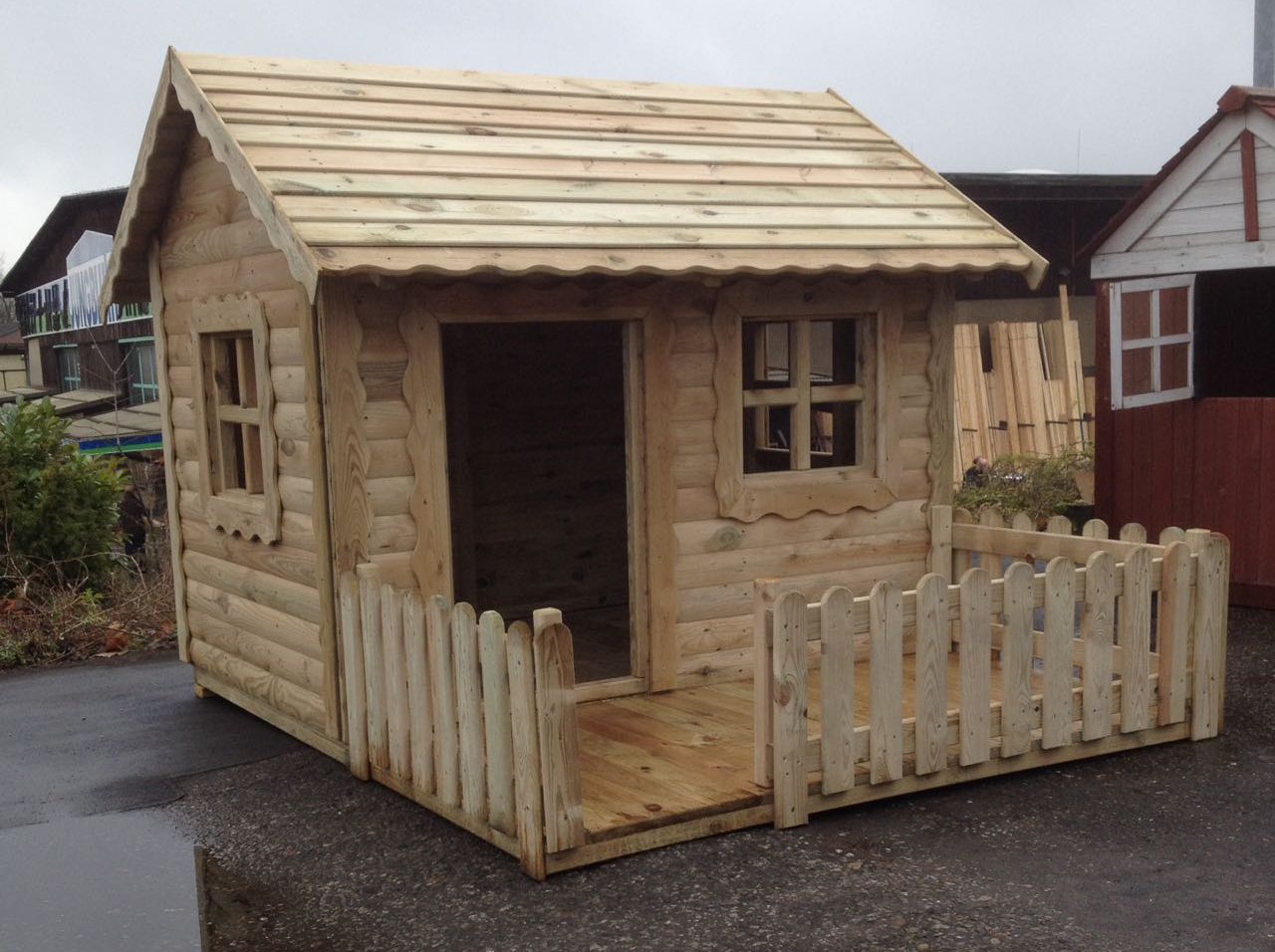 Kinderspielhaus Diana aus hellem Holz, Maße 170 x 220 x 155 cm, kesseldruckimprägniert, mit Satteldach, zwei Fenstern, offenem Eingang und kleiner umlaufender Veranda mit Holzzaun, im Außenbereich aufgestellt