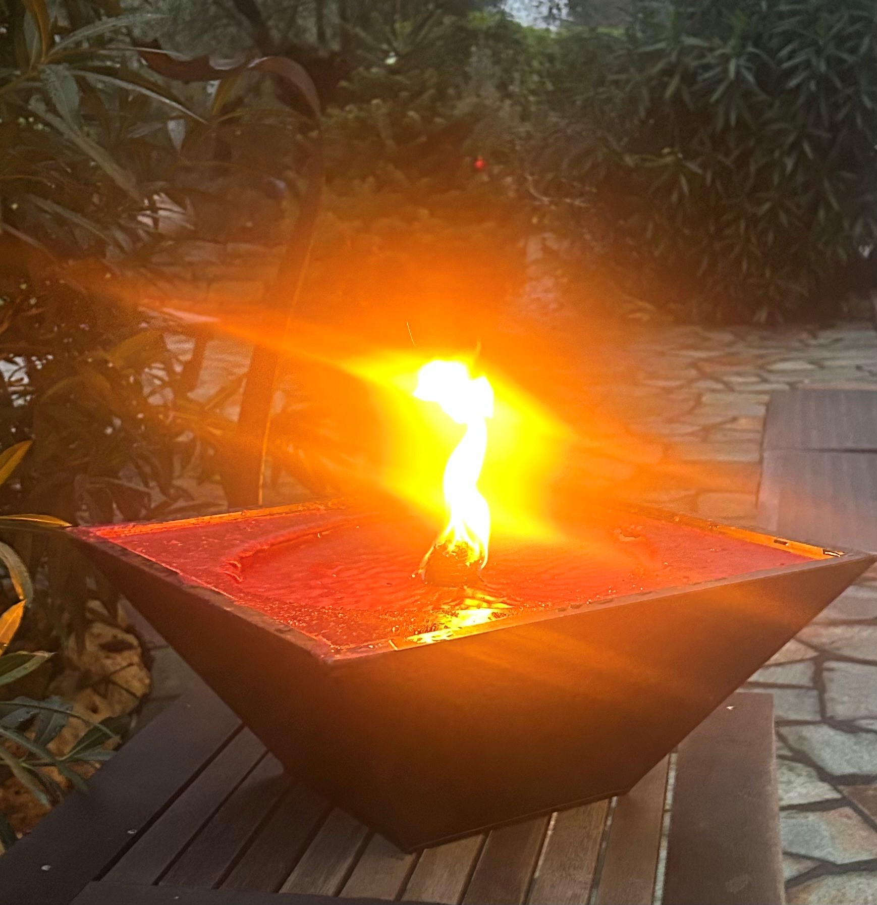 Rote Wachsschale mit brennender Flamme in einem schwarzen, geometrischen Behälter im Freien auf einem Holztisch, umgeben von Pflanzen und Steinboden.