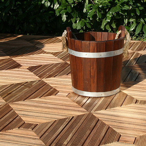 Holzeimer mit Metallbändern und Taugriffen auf geometrisch gemusterten Holzfliesen-Terrasse, teilweise im Sonnenlicht, grüner Busch im Hintergrund.